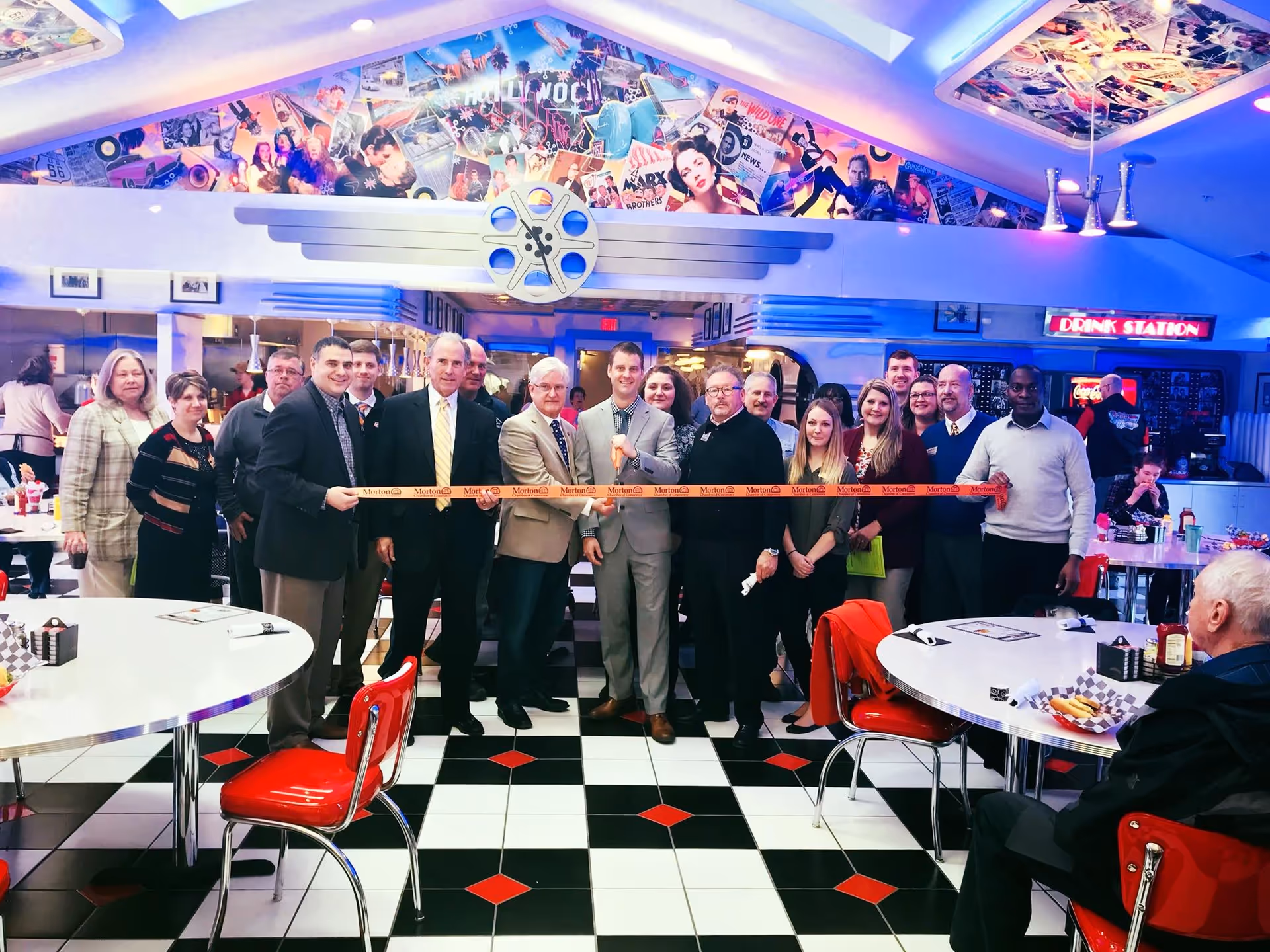 A group of people standing in a retro-themed diner with black and white checkered floor and red chairs, holding a ribbon for a ribbon-cutting ceremony. The diner has colorful movie-themed wall art and a drink station in the background.