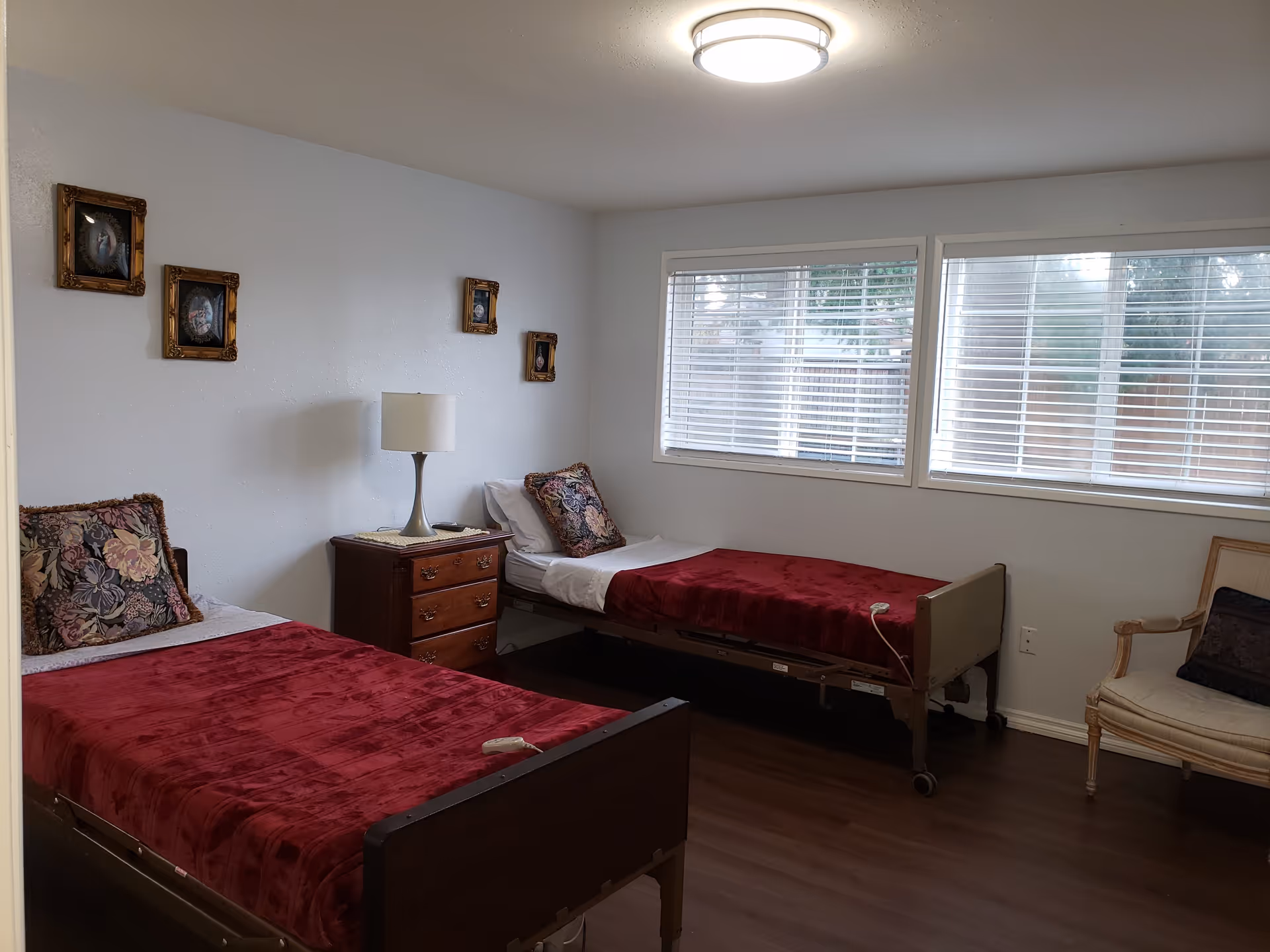 Two single beds with red covers in a simple room with a nightstand, lamp, framed pictures on the wall, and large windows.