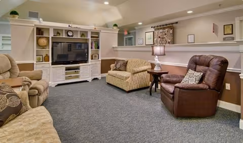 A cozy living room area with beige and brown tones featuring a large entertainment center with a TV, shelves with decorative items, a beige sofa, a patterned loveseat, a brown leather recliner, a side table with a lamp, and carpeted flooring.