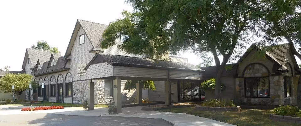 Exterior front view of a senior living building with peaked roofs, stone-and-siding facade, a covered porte-cochere driveway, trees, and landscaping.