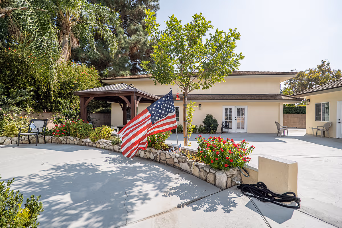 Outdoor courtyard area of an assisted living facility with a small tree, American flag, flower beds, patio chairs, and a wooden gazebo. The building has light-colored walls and multiple doors leading outside.
