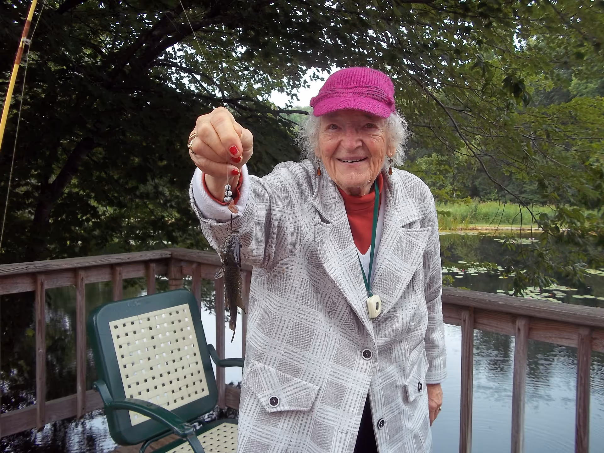 An elderly woman wearing a purple cap and a light-colored plaid coat is standing on a wooden deck by a pond, holding up a small fish she caught with a fishing rod. There is a green chair with a woven seat and backrest next to her, and trees and water lilies are visible in the background.
