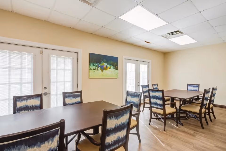 A bright dining room with two rectangular dark wood tables surrounded by chairs with patterned blue and white backs and beige seats. The room has light beige walls, wood flooring, two sets of French doors with blinds, and a colorful fish painting on the wall.