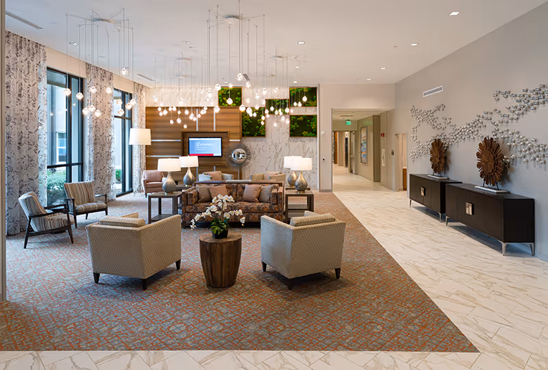 Bright, modern senior living lounge with sofas and chairs arranged around a coffee table, decorative pendant lights, and a hallway entrance.