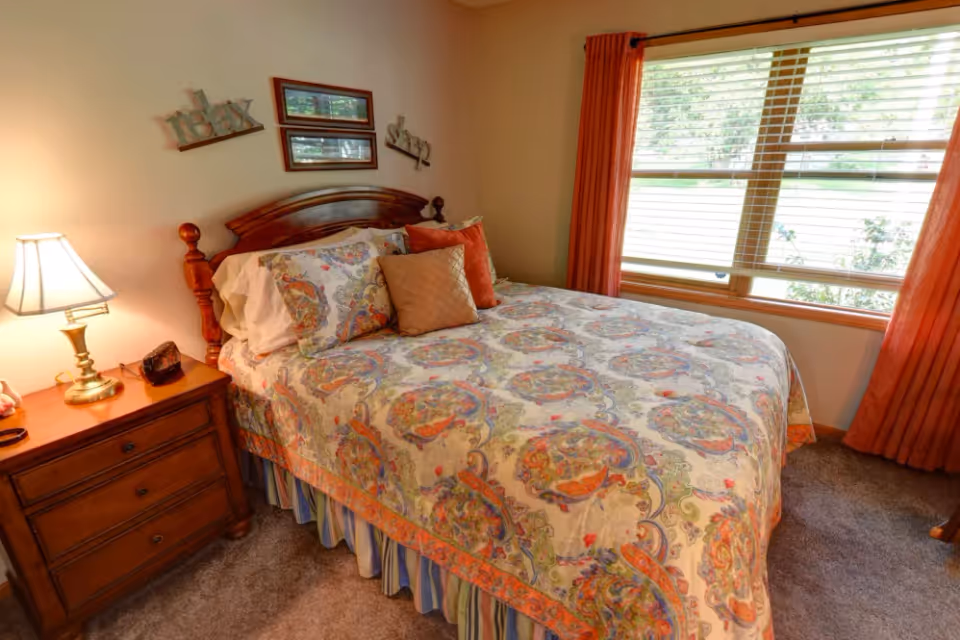 A cozy bedroom with a wooden bed frame and a colorful patterned bedspread. There are three decorative pillows on the bed. A wooden nightstand with a lamp and some small items is beside the bed. The room has a large window with orange curtains and blinds, letting in natural light.