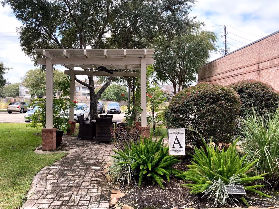 Outdoor garden area with a brick pathway leading to a white pergola with ceiling fans and wicker chairs underneath. Surrounding the pathway are green plants, bushes, and trees. A sign with the letter 'A' is placed among the plants. In the background, there is a parking lot with several cars and a brick building wall on the right side.