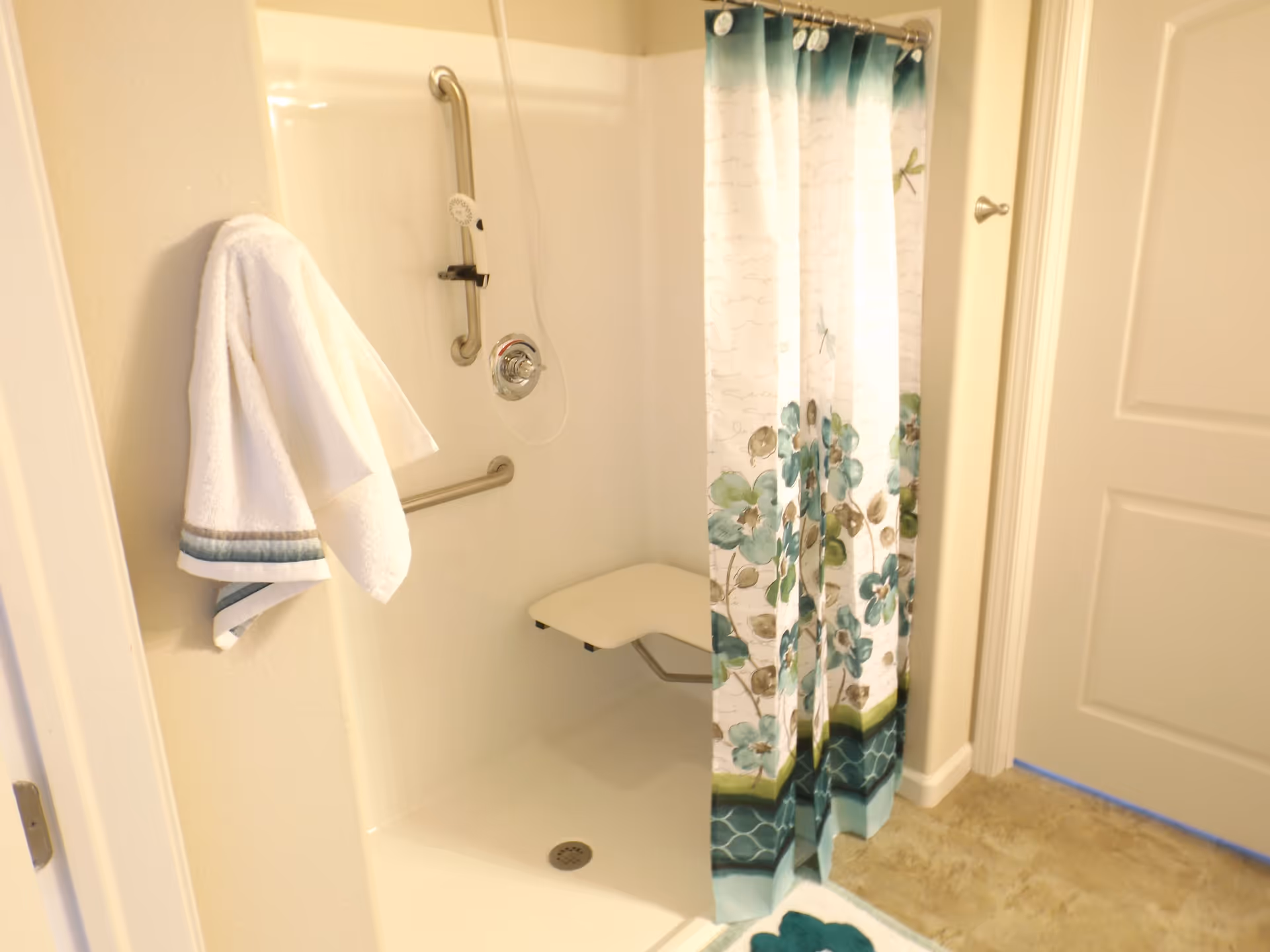 A bathroom shower area with a white wall-mounted foldable seat, grab bars, a handheld showerhead, and a floral-patterned shower curtain in shades of teal, green, and white. A white towel with teal stripes hangs on a hook on the wall next to the shower.
