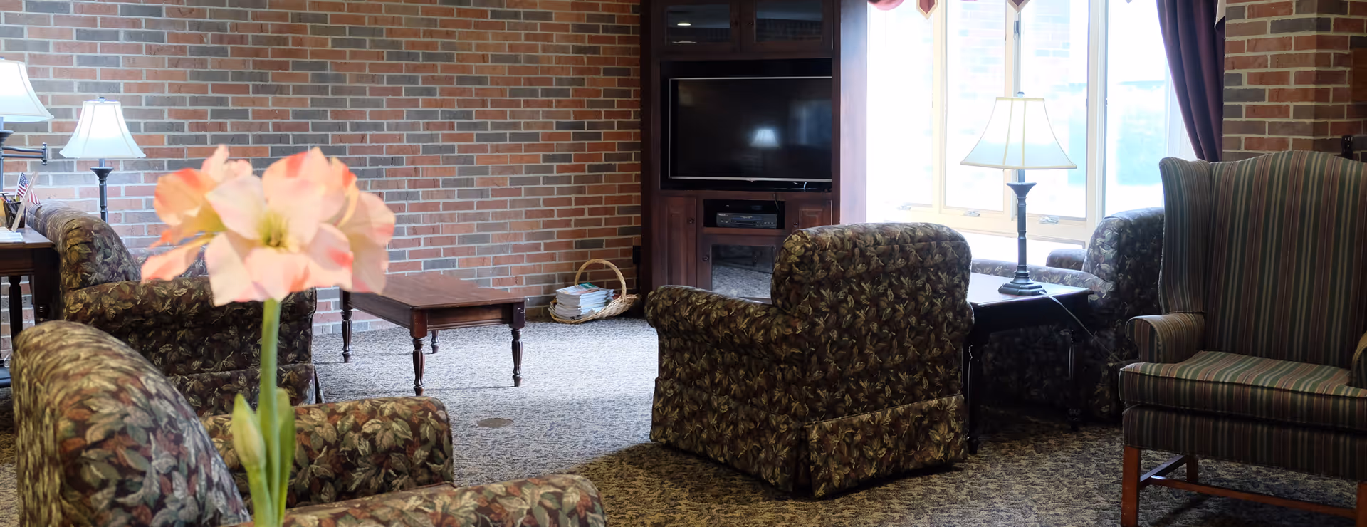 A cozy assisted-living lounge with patterned armchairs, a coffee table and TV against a brick wall, lamps by a window, and a pink flower in the foreground.