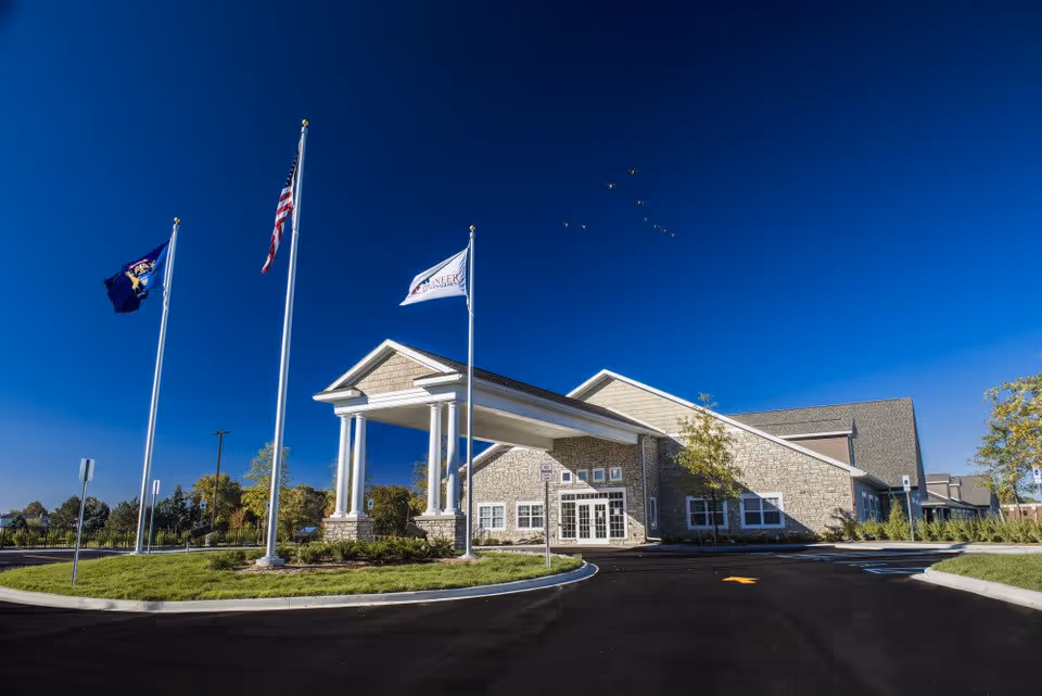 Exterior view of Lakeside Manor senior living facility with a clear blue sky, three flagpoles displaying flags, and a paved driveway leading to the entrance with white columns.