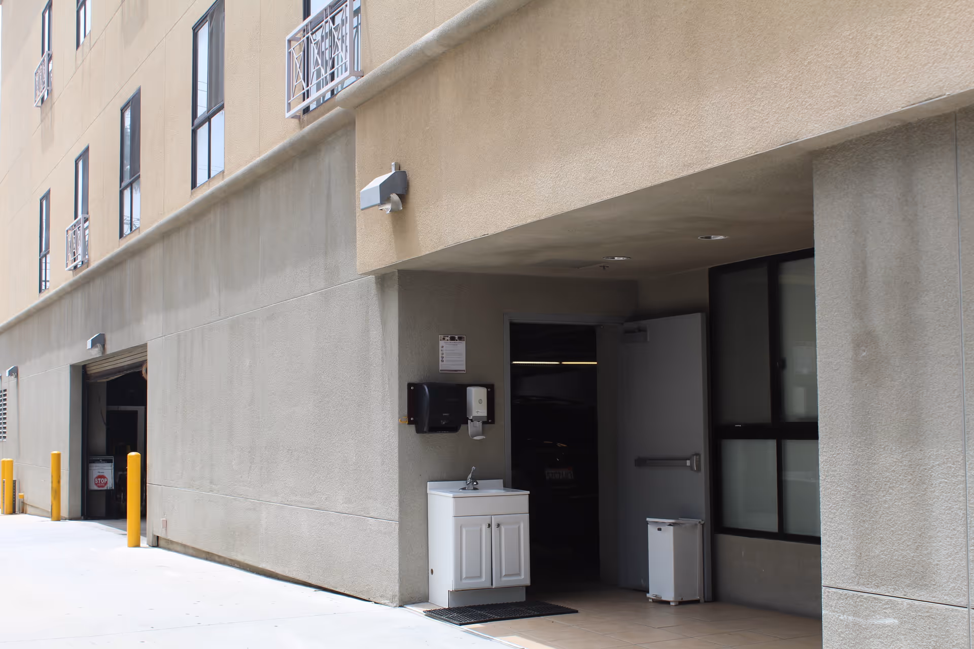 Exterior view of a beige building with a small white sink and soap dispenser mounted on the wall outside near an open door leading inside. There are yellow bollards along the side of the building and a partially visible garage door with a stop sign inside.