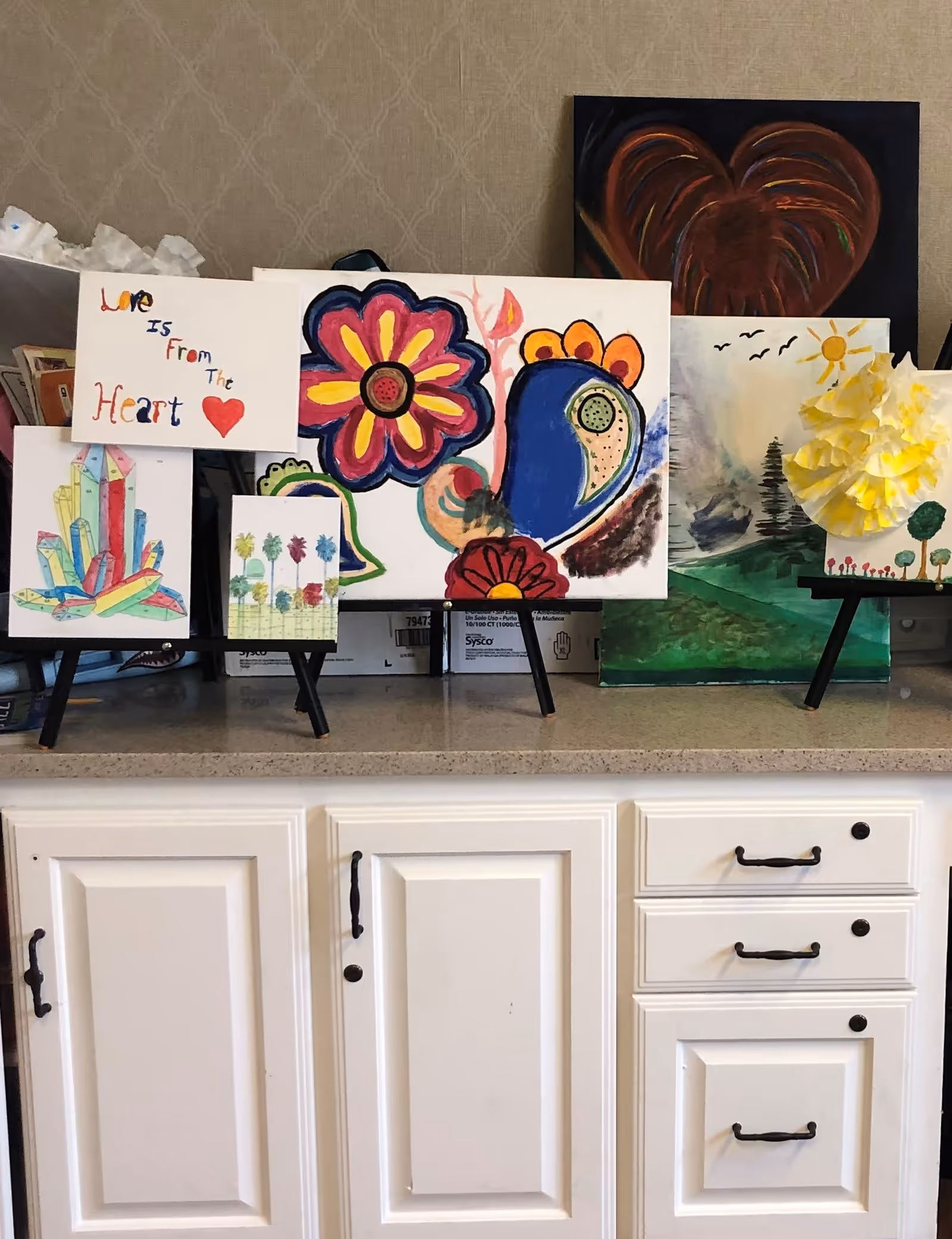 A countertop with several colorful paintings displayed on small easels. The paintings include a large flower, a heart, a cluster of crystals, palm trees, a landscape with trees and birds, and a yellow paper flower. Below the countertop are white cabinets with black handles.