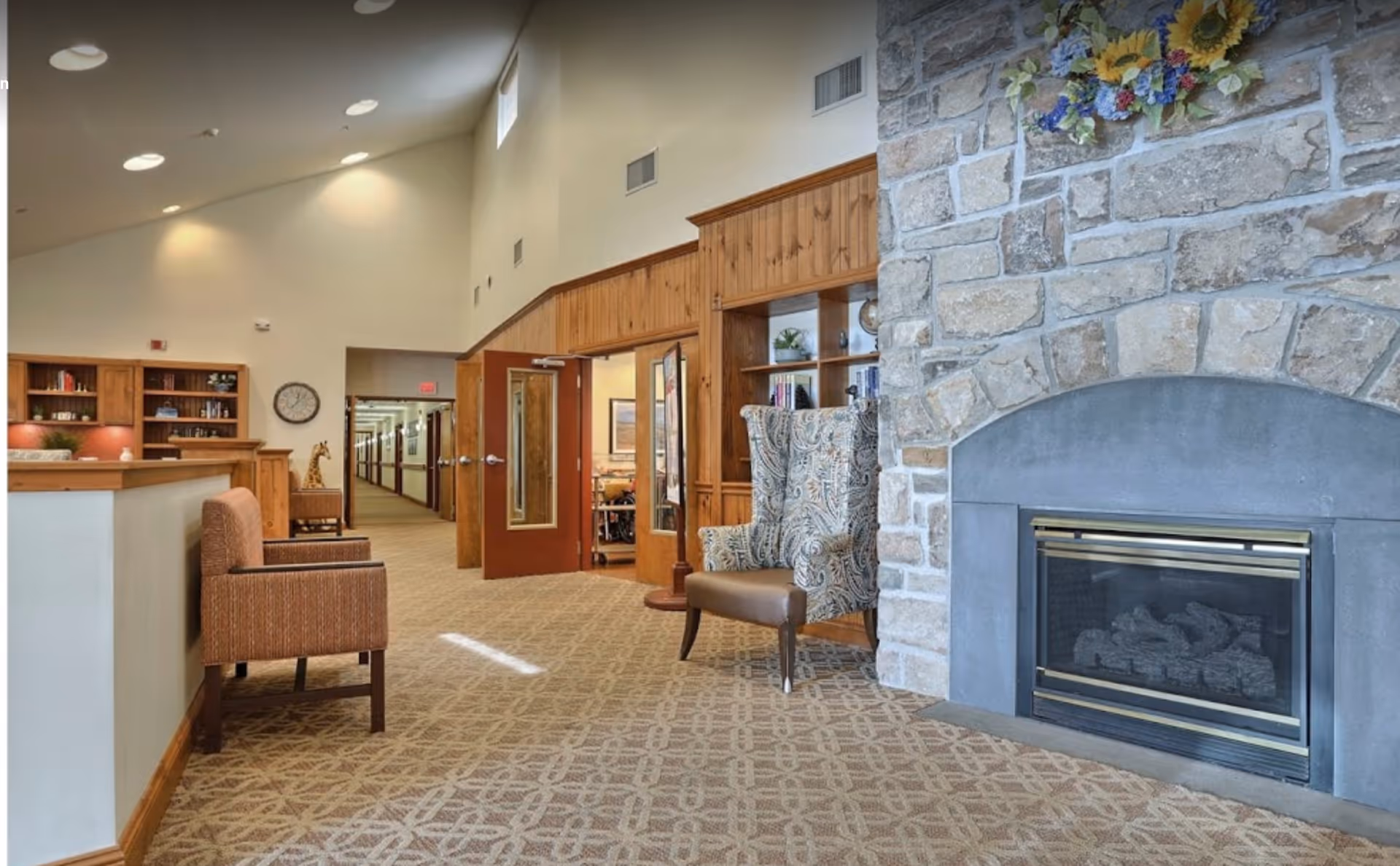 Carpeted senior living lounge with a stone fireplace, patterned armchairs, bookshelves and a hallway leading to rooms.