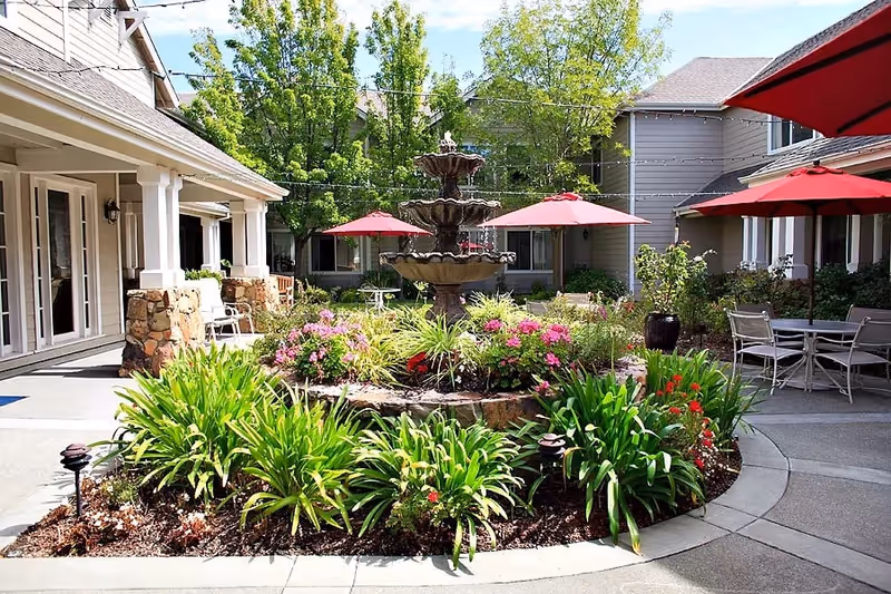 Outdoor courtyard area with a multi-tiered water fountain surrounded by lush green plants and colorful flowers. Several red patio umbrellas shade tables and chairs arranged around the courtyard. The surrounding buildings have beige siding and stone accents, with large windows and doors opening to the courtyard.