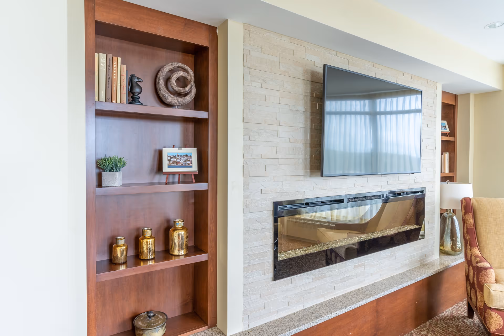 A cozy living room area featuring a mounted flat-screen TV above a modern electric fireplace set in a light stone wall. To the left, there is a wooden built-in bookshelf with decorative items including books, a small plant, a framed picture, and gold-colored vases. A beige armchair with a patterned red and gold cushion is partially visible on the right side.