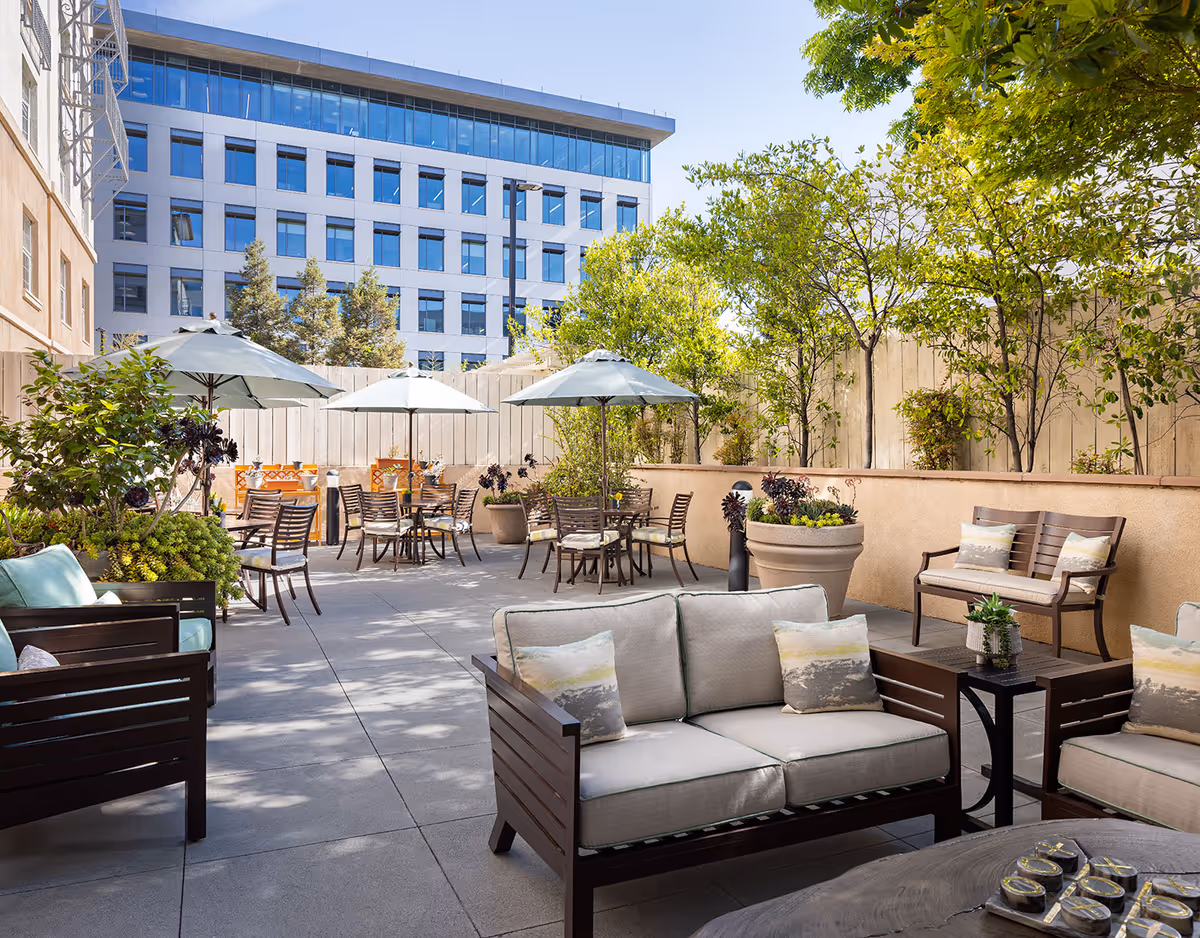 Outdoor lounge patio with sofas, chairs, tables, umbrellas, potted plants, and trees beside a modern building.