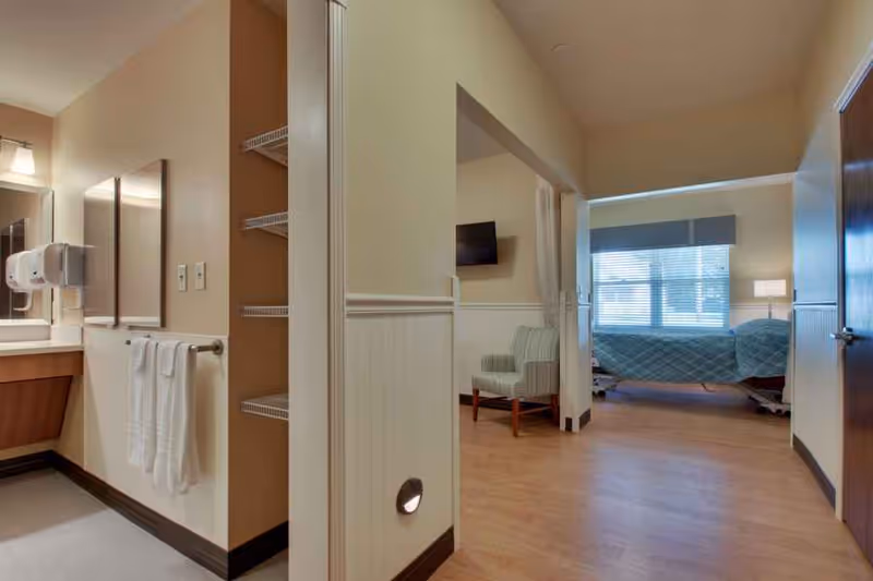 View of a senior care facility room showing a hospital-style bed with a blue quilt, a window with blinds, a striped armchair, and a wall-mounted TV. The image also shows part of a bathroom area with a sink, mirror, towel rack with white towels, and shelves.
