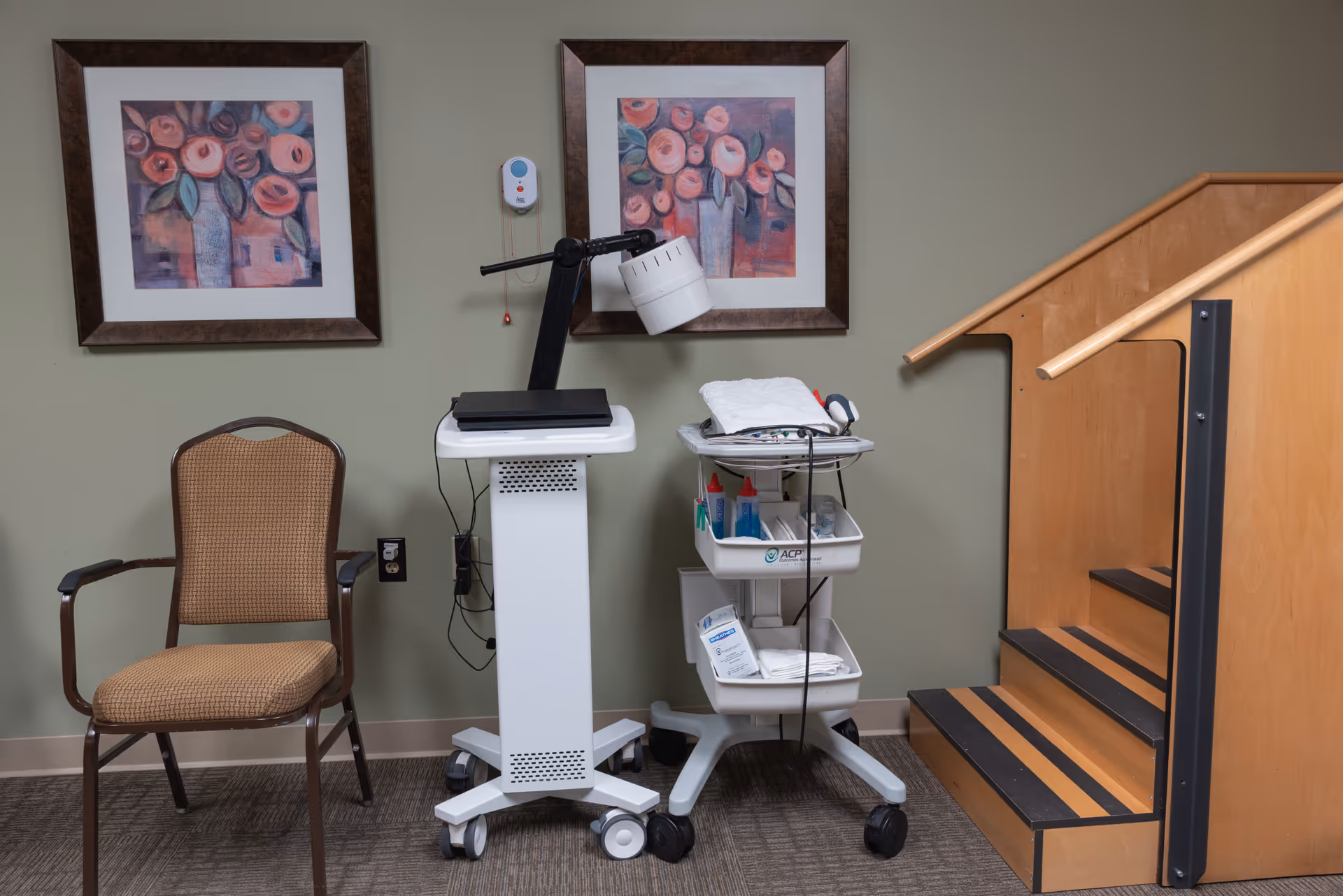 A corner of a room with a brown cushioned chair, a white medical equipment cart with supplies, a white stand with a lamp, two framed floral paintings on a green wall, and a wooden staircase with black anti-slip strips.