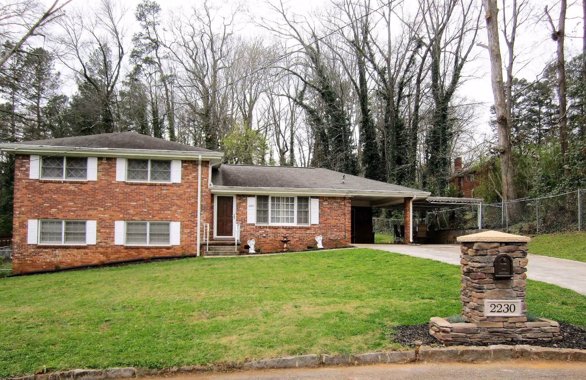 Front exterior view of a brick house with white window shutters, a driveway leading to a covered carport, a green lawn, and a stone mailbox with the number 2230. Tall trees without leaves are visible in the background.