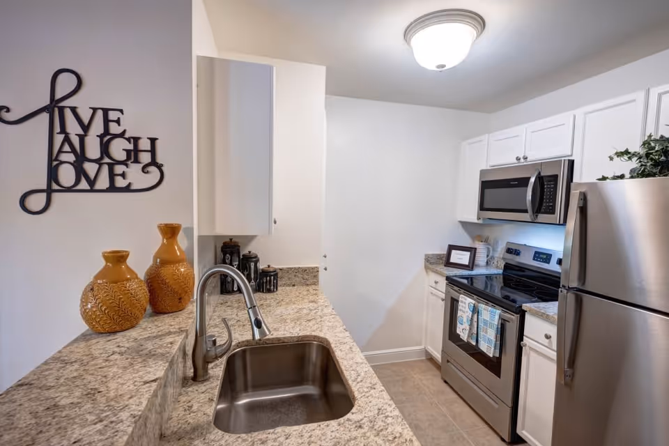 A modern kitchen with granite countertops, a stainless steel sink, stainless steel refrigerator, stove, and microwave. On the left wall, there is a decorative metal wall art with the words 'Live Laugh Love'. Two orange decorative vases are placed on the countertop near the sink.
