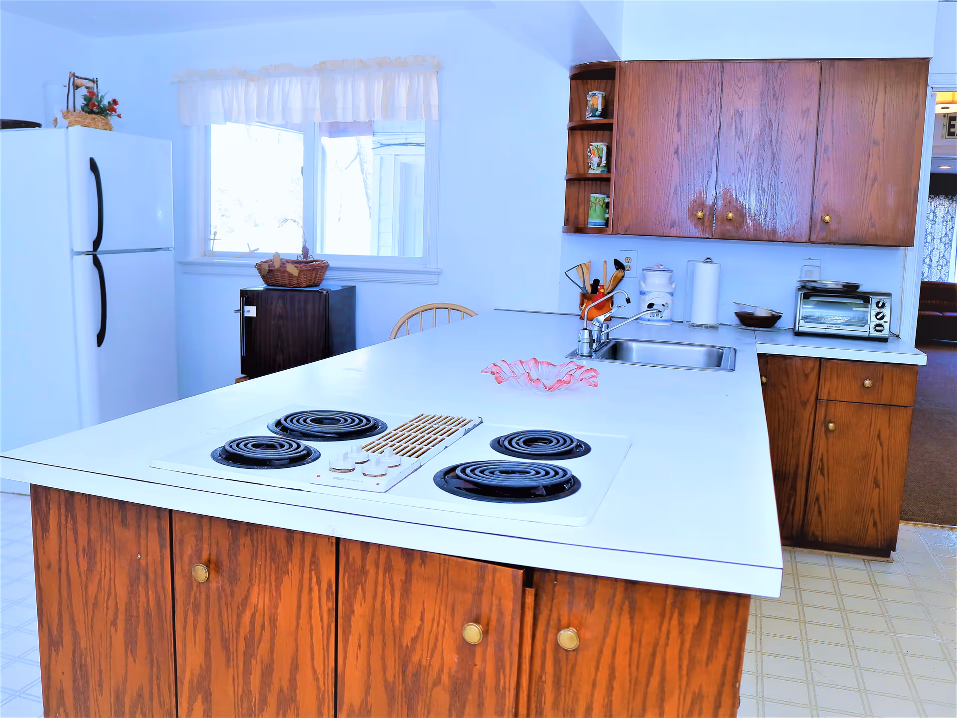 Kitchen with a large white island containing an electric stovetop and sink, wood cabinets, refrigerator, and a window.