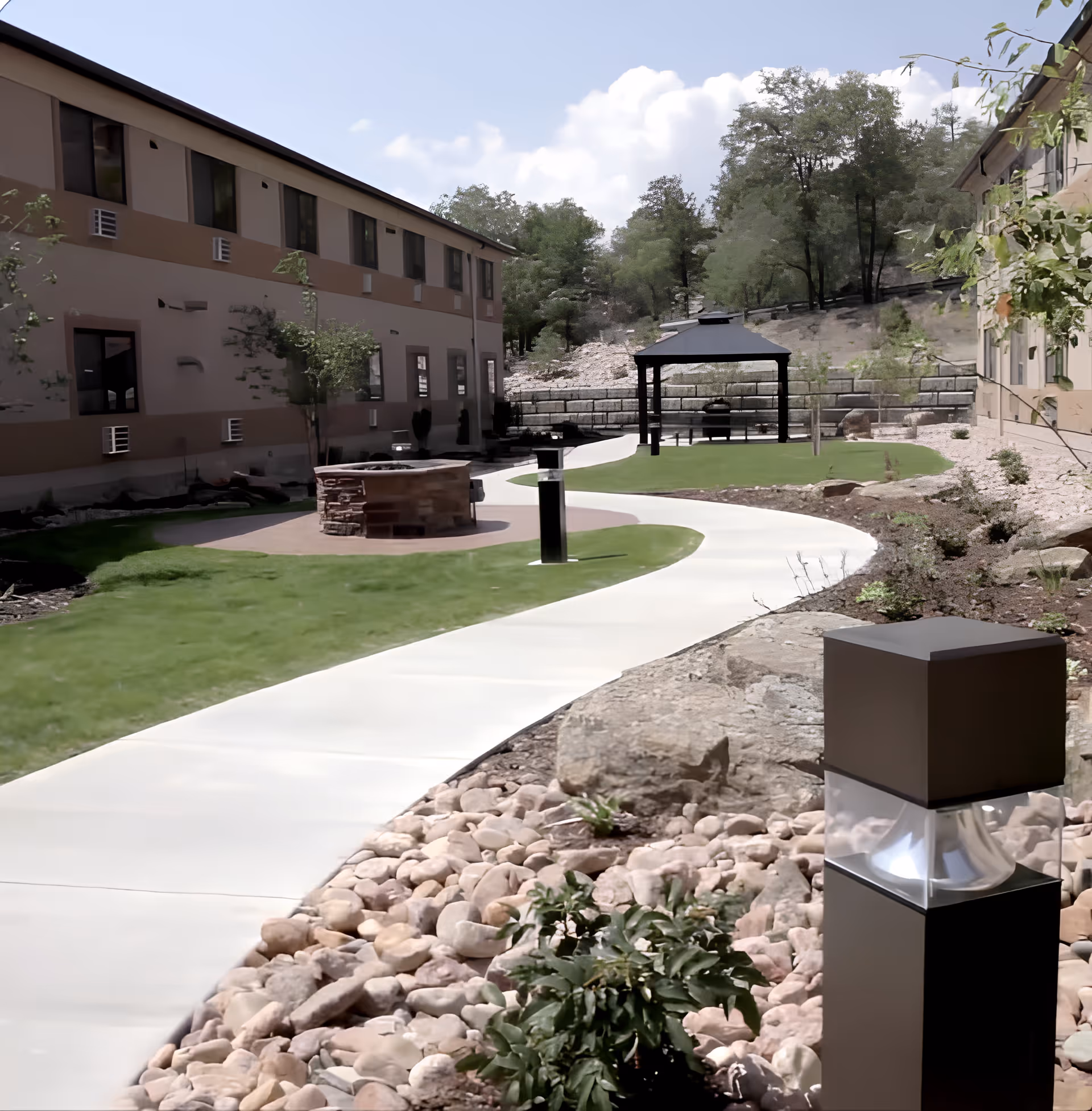 Outdoor garden area at Point of the Pines Gardens featuring a winding concrete pathway, green grass patches, a stone fire pit, a gazebo with seating, and surrounding trees and shrubs. Two beige two-story buildings flank the garden on either side.