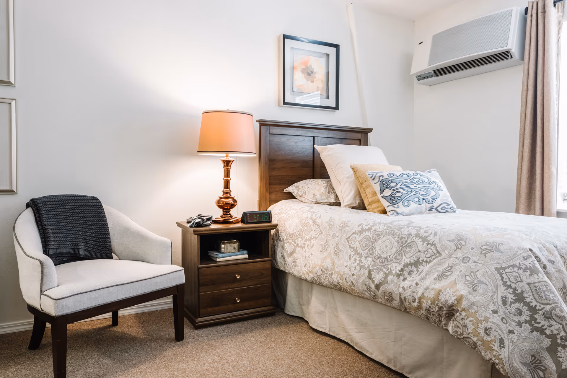 A tidy bedroom with a single bed, patterned bedding, a wooden nightstand and lamp, and an upholstered chair.