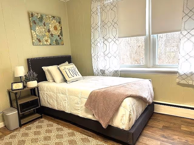 A bright and cozy bedroom with a single bed featuring white bedding, a decorative pillow with the word 'LOVE', and a light pink throw blanket. The room has a wooden floor, a patterned area rug, a small bedside table with a lamp and a plant, and a large window with sheer patterned curtains letting in natural light. A floral artwork hangs on the wall above the bed.