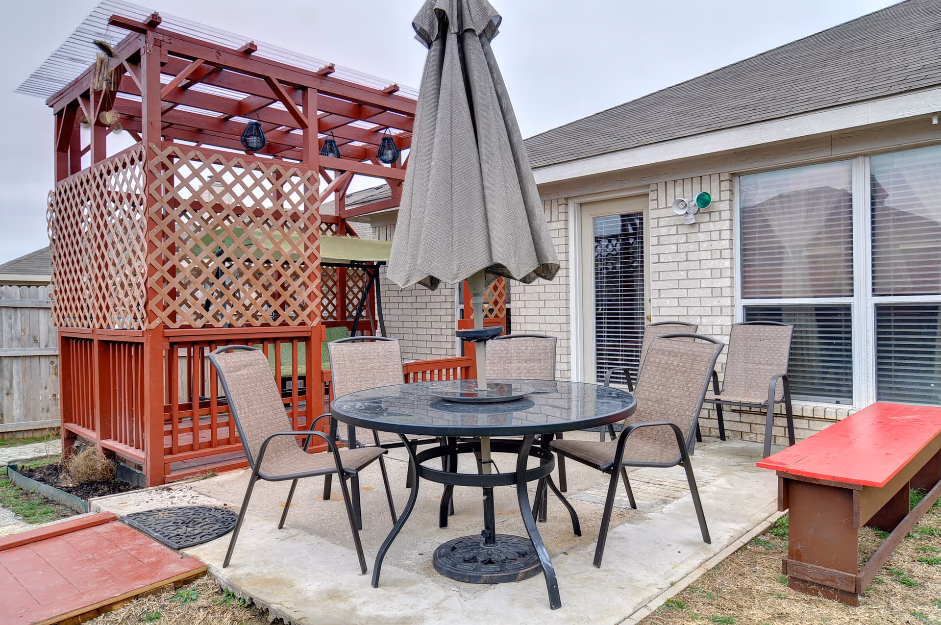 Outdoor patio with a round glass table, umbrella and chairs beside a red lattice pergola and brick building.