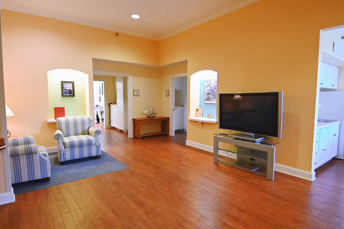 Spacious living room with wood floors, two striped armchairs, a flat-screen TV on a stand, and warm yellow walls.