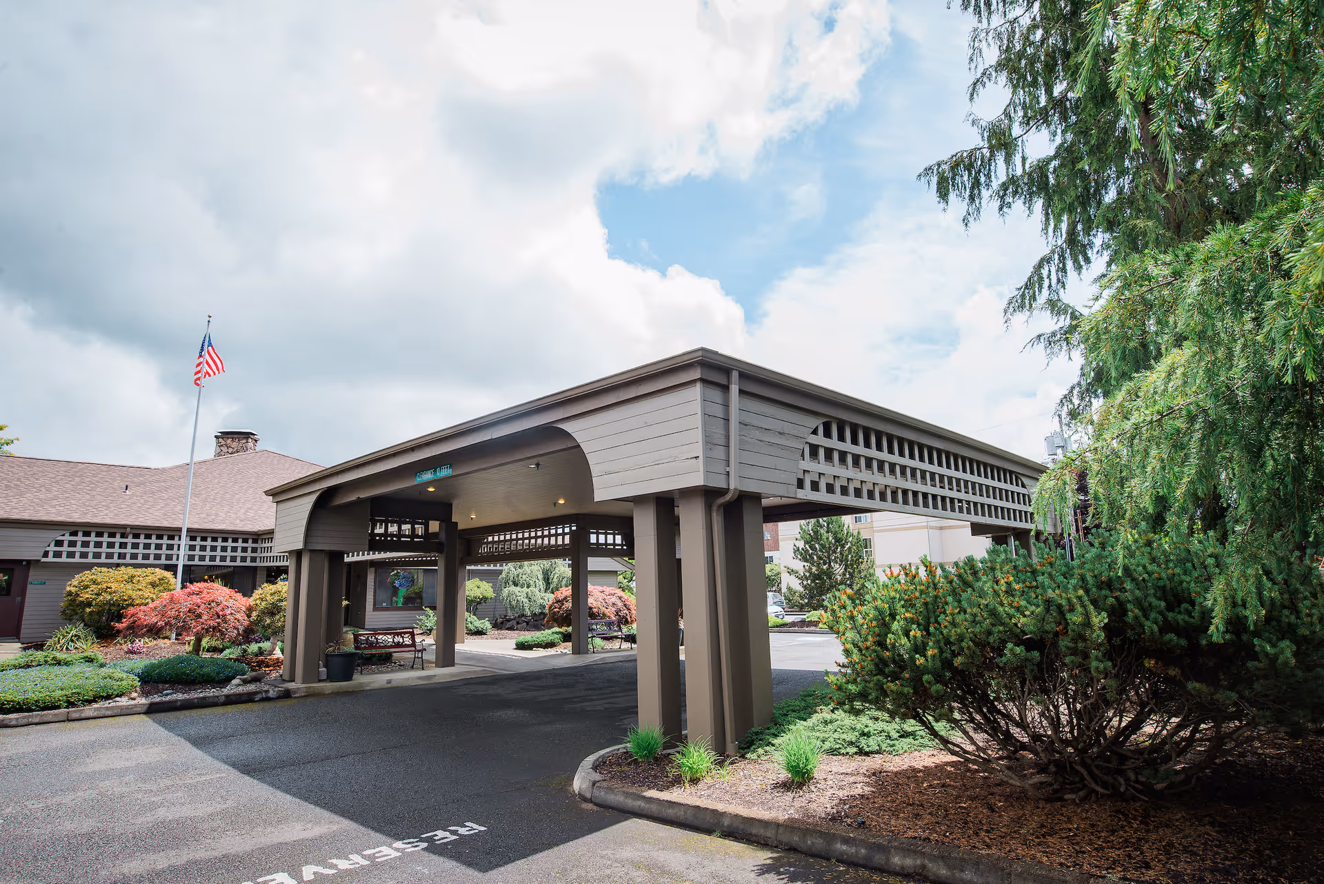 Entrance area of Channel Point Village senior living facility with a covered drop-off zone, landscaped bushes and trees, an American flag on a flagpole, and partly cloudy sky.
