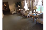 A cozy living room area with several upholstered armchairs and small wooden tables arranged along a window with floral curtains. There are two table lamps and a potted plant in the corner, with a carpeted floor and a hallway visible in the background.