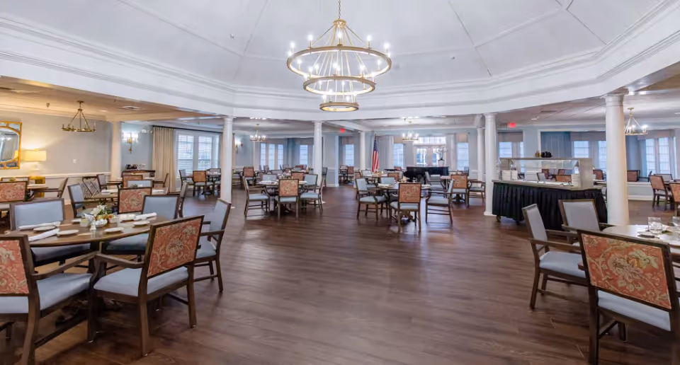 Spacious dining room with multiple tables and chairs arranged neatly on a wooden floor. The room features elegant chandeliers hanging from a white coffered ceiling, large windows with curtains, and decorative columns. Tables are set with plates, napkins, and small floral centerpieces.
