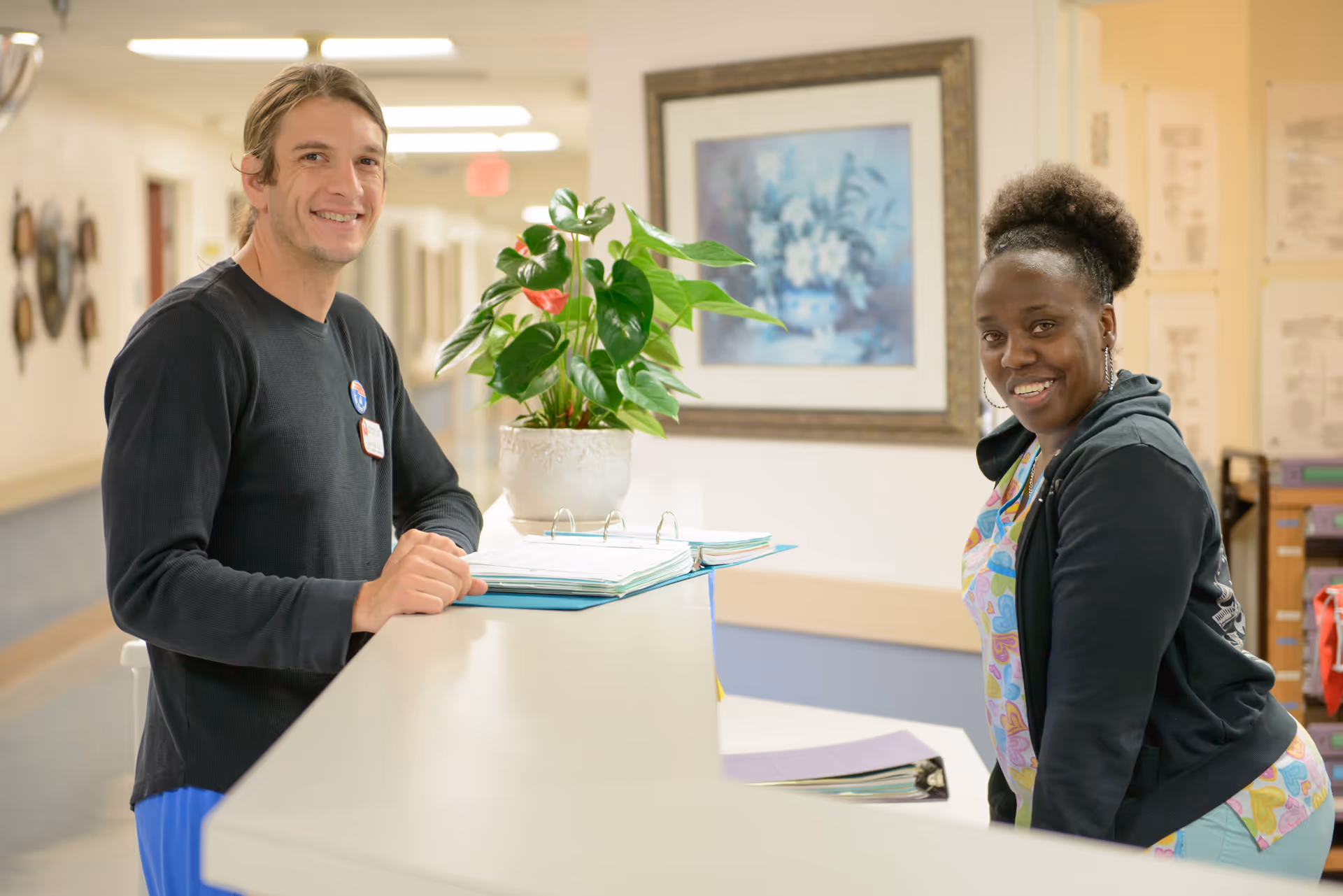 Two healthcare workers smiling and standing at a reception desk in a well-lit hallway of a senior care facility. One is a man wearing a black long-sleeve shirt and blue pants, and the other is a woman wearing colorful scrubs and a black jacket. A potted plant and open binders are on the desk, with framed artwork and documents on the walls behind them.