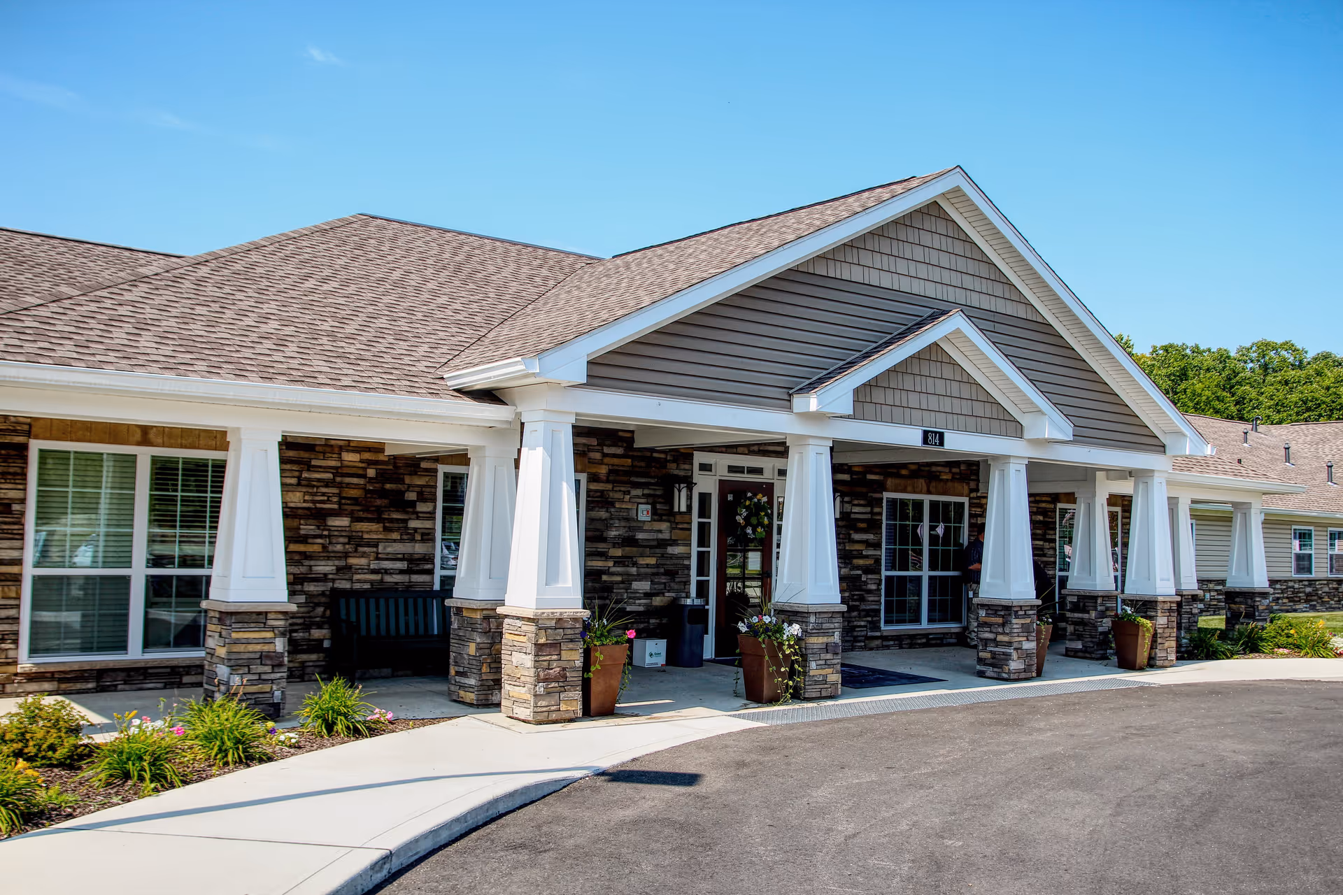 Exterior front view of a senior living facility building with stone and siding facade, white columns supporting a covered entrance, large windows, and a paved driveway under a clear blue sky.
