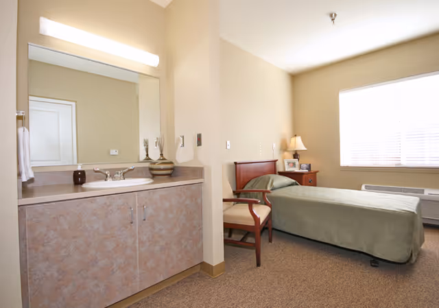 A single bedroom in an assisted living facility featuring a bed with a green bedspread, a wooden chair, a nightstand with a lamp and framed photos, a large window with blinds, and a sink with a mirror and countertop in the corner.