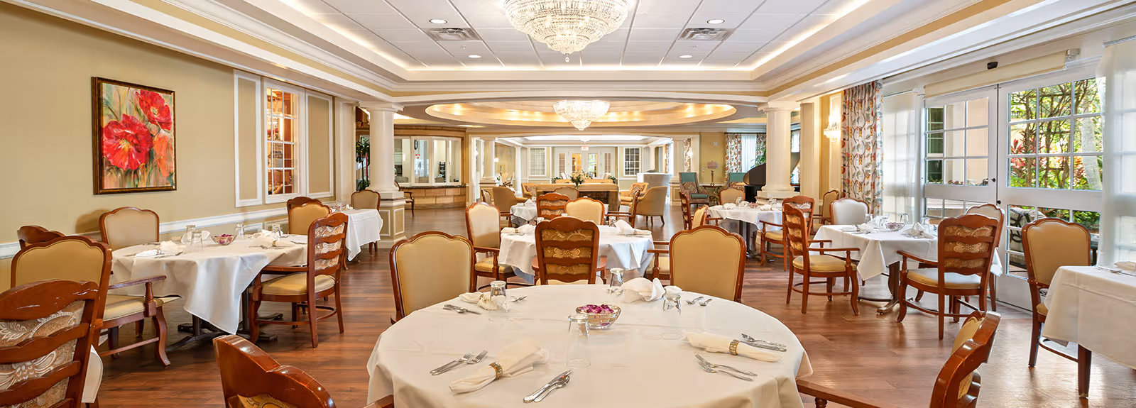 Elegant senior living dining room with round tables set with white linens, wooden chairs, chandeliers and large windows.