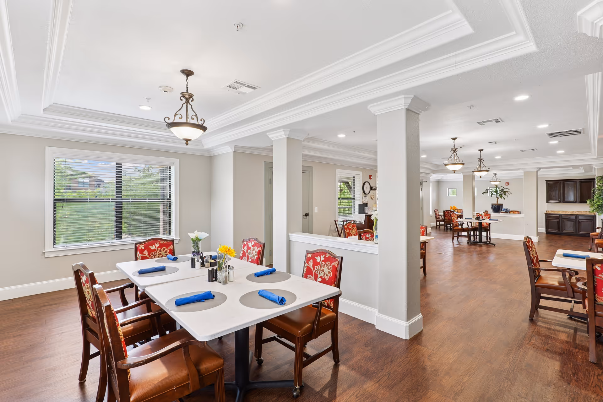 Bright and spacious dining area in a senior living facility with multiple tables and chairs. The tables are set with blue napkins and small flower vases. Large windows allow natural light to fill the room, and decorative ceiling lights hang above. The floor is wooden, and the walls are painted light beige with white trim.