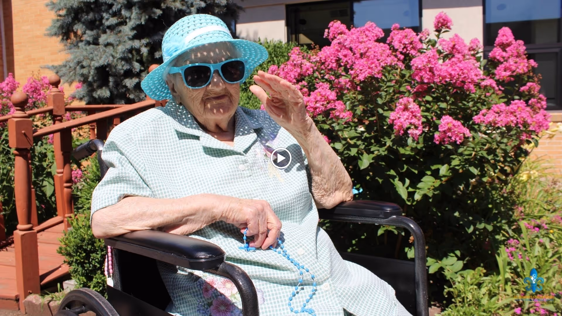 An elderly woman wearing a light blue hat and matching sunglasses sits in a wheelchair outdoors in front of a bush with bright pink flowers. She is dressed in a light green checkered shirt and holds a blue beaded necklace in her hand while waving with the other hand. The background includes a wooden railing and a building with windows.