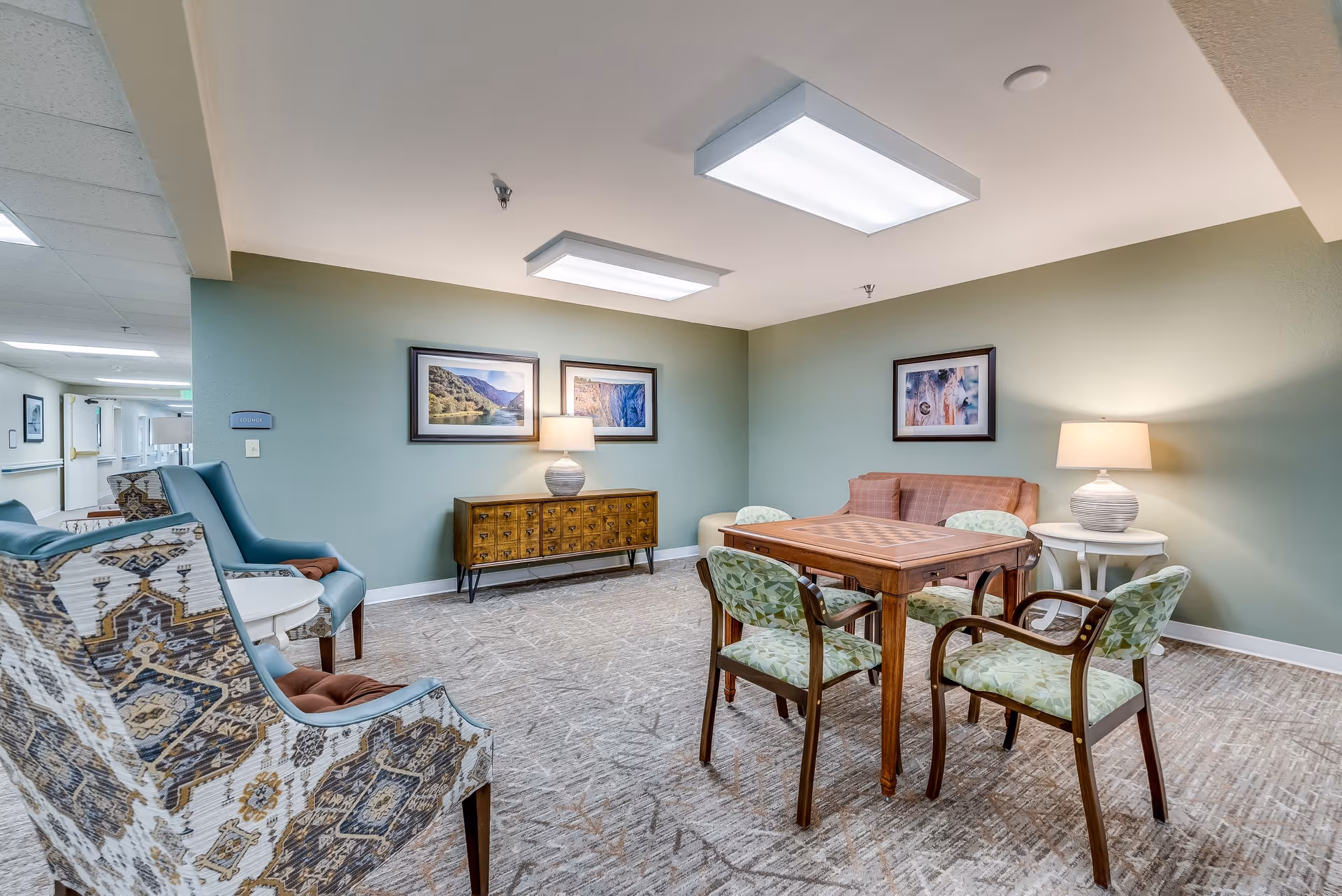 A comfortable senior living common room with a wooden game table and chairs, upholstered armchairs and sofas, lamps, and framed artwork on the walls.