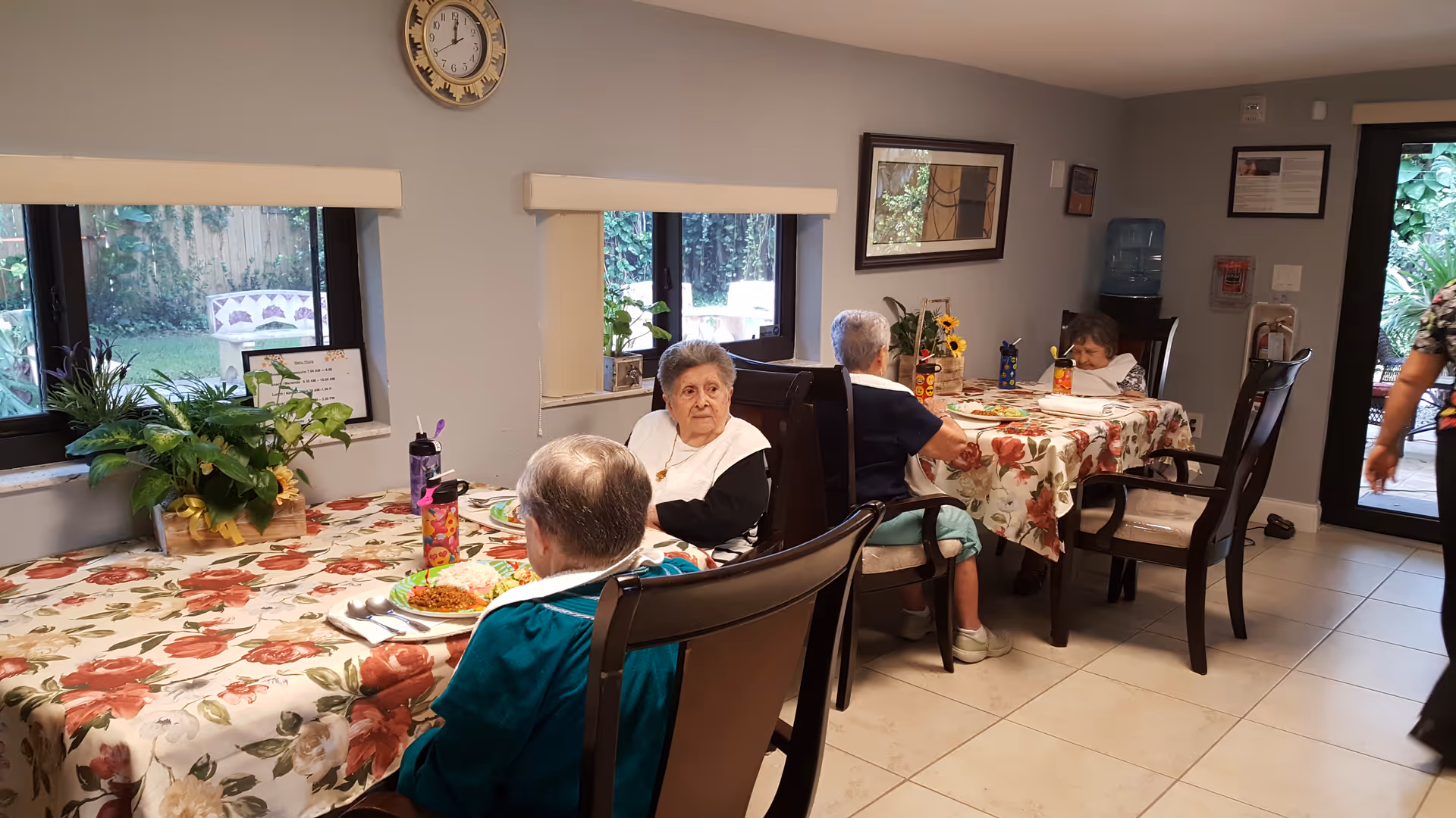 A dining room in a senior living facility with three elderly women seated at two tables covered with floral tablecloths. Plates of food and water bottles are on the tables. Large windows show greenery outside, and a clock is mounted on the wall above the windows. A water dispenser and framed pictures are visible on the walls.