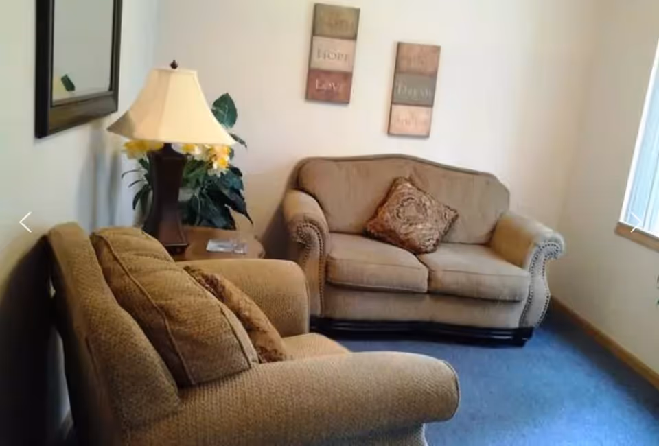 A cozy living room with two beige upholstered sofas, one with a decorative pillow. There is a wooden side table between the sofas with a lamp and a plant. Two framed wall art pieces with inspirational words hang on the wall above the sofa. A window with blinds allows natural light into the room.