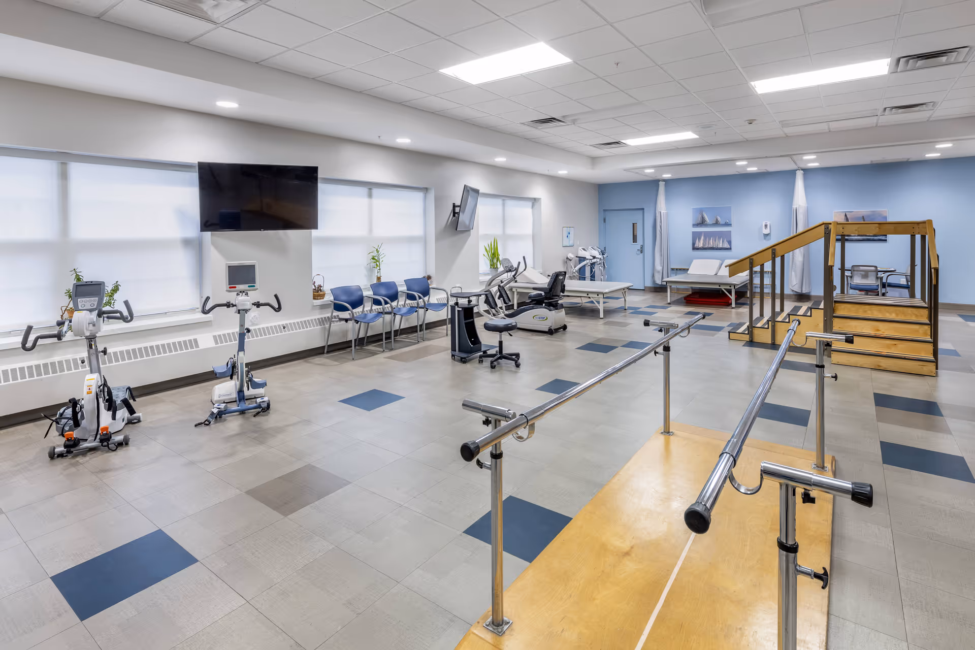 A spacious physical therapy room with exercise bikes, parallel bars for walking practice, a wooden staircase for rehabilitation, several chairs lined up against the wall, and treatment tables. The room has large windows with blinds, light blue walls, and ceiling lights.