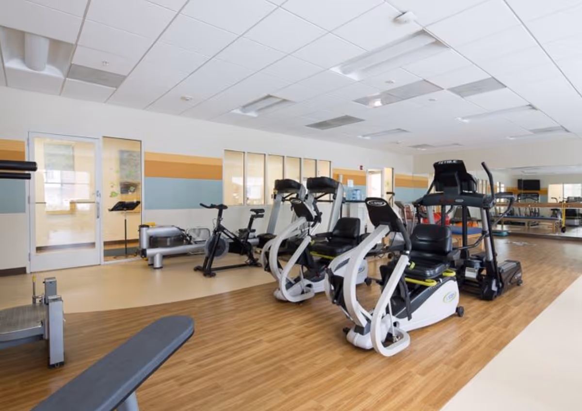 A fitness room in a senior living facility featuring exercise equipment including stationary bikes, treadmills, and weight machines. The room has wood and beige flooring, large windows, and a mirrored wall.