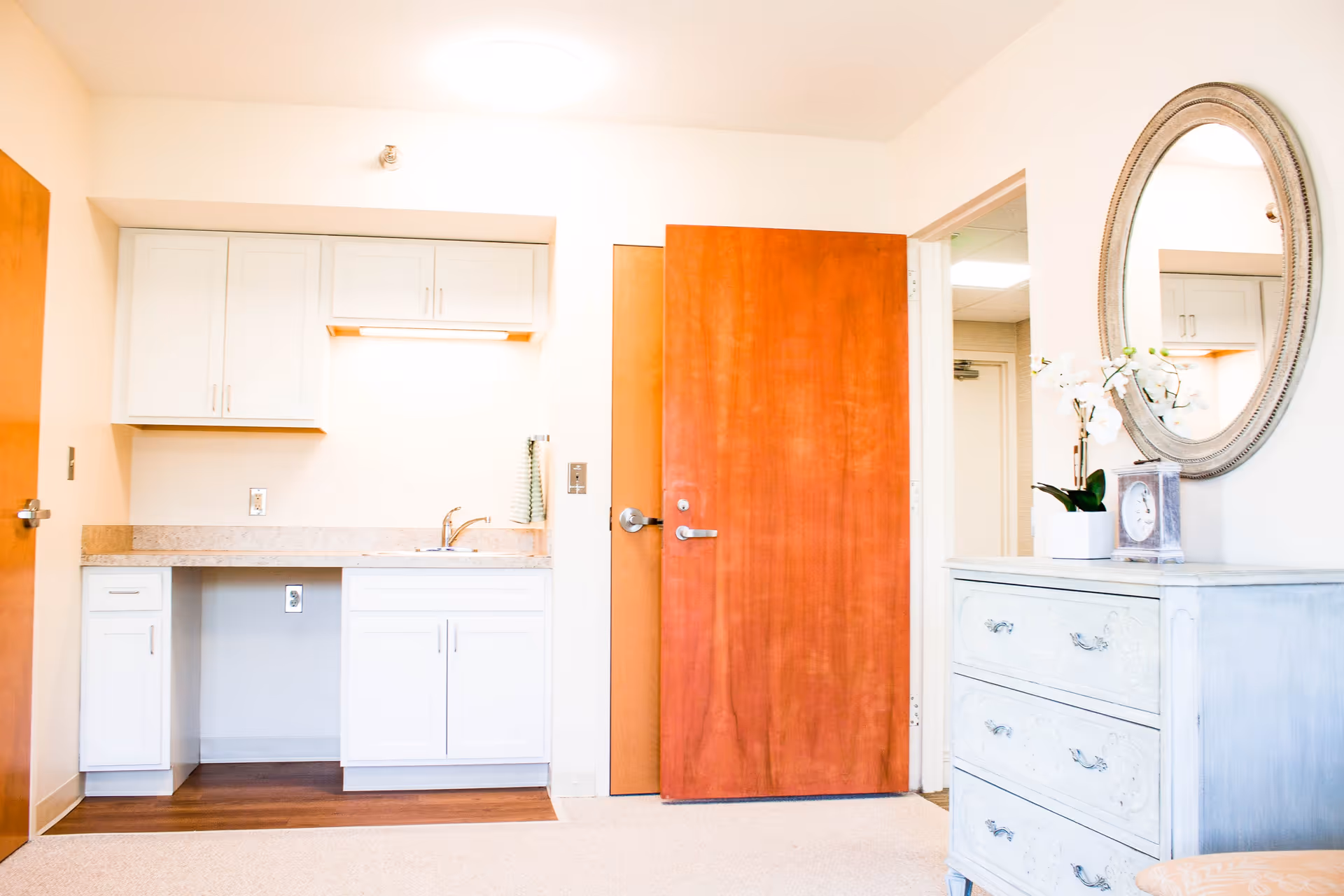 A bright room featuring a small kitchenette with white cabinets, a countertop, and a sink. To the right, there is a wooden door and a light-colored dresser with a round mirror above it. A small potted plant and a clock are placed on the dresser.