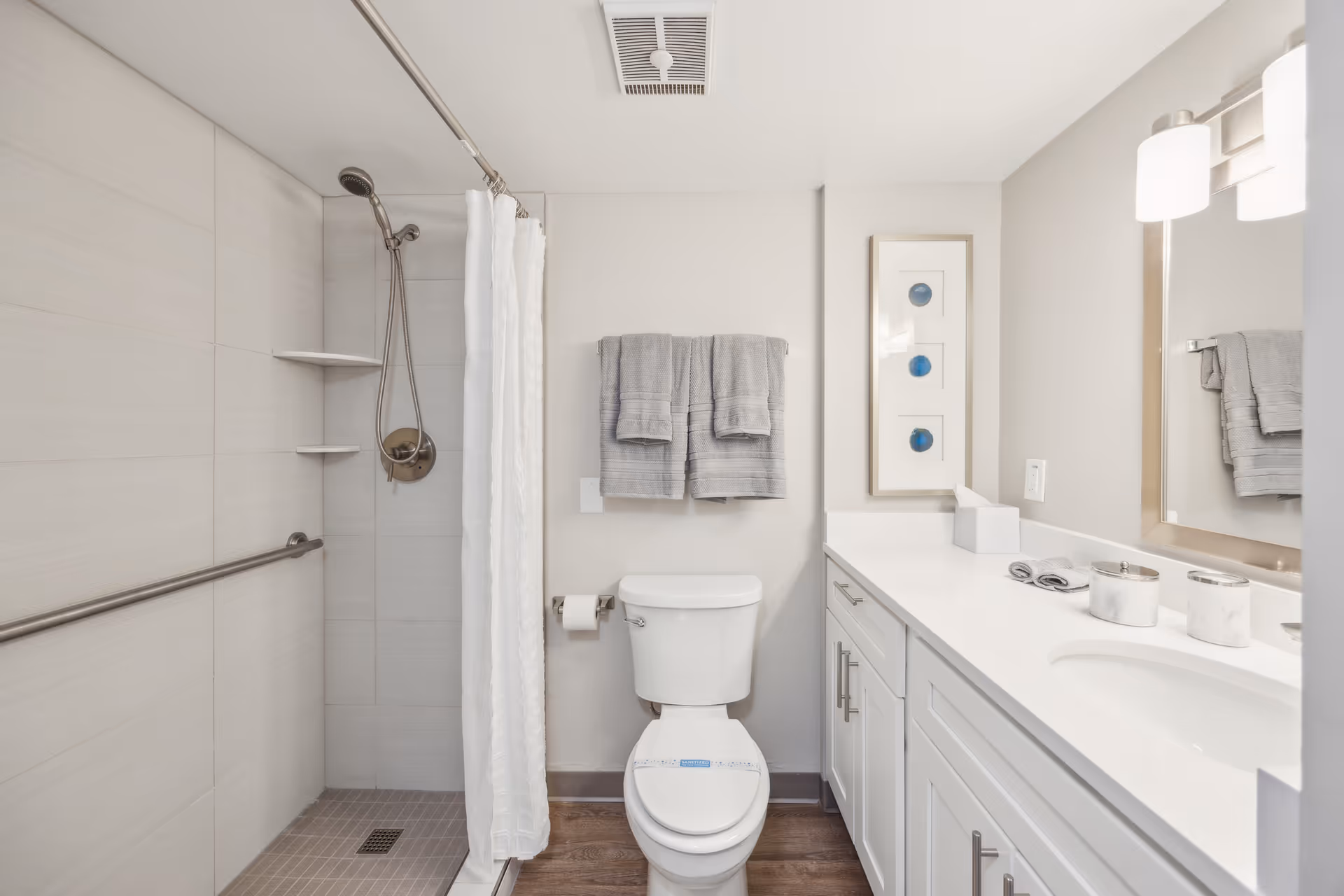 A clean and modern bathroom featuring a walk-in shower with a handheld showerhead and white curtain, a white toilet with a sanitized seal, gray towels hanging on a rack above the toilet, and a white vanity with a sink, mirror, and light fixtures. The walls are light-colored, and the floor has wood-like tiles.