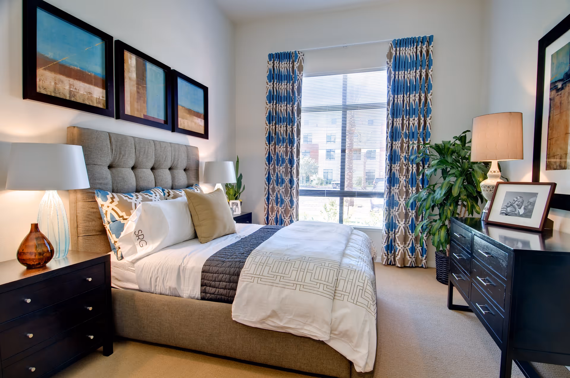 A well-lit bedroom with a beige upholstered bed featuring white and patterned bedding. There are two dark wooden nightstands with lamps on either side of the bed. Above the bed are three framed abstract paintings. A large window with blue and beige patterned curtains lets in natural light. On the right side, there is a dark wooden dresser with a lamp, a framed black and white photo, and a green potted plant beside it.