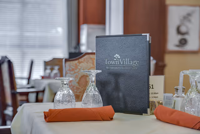 A dining table set with two upside-down wine glasses, an orange cloth napkin, and a black menu folder labeled 'Town Village Retirement Community' in a warmly lit dining room with wooden chairs and large windows with blinds in the background.