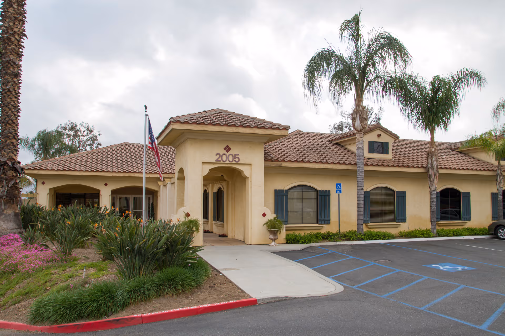 Exterior view of a single-story building with a tiled roof and beige stucco walls, featuring the number 2005 above the entrance. There are palm trees and landscaped greenery around the building, an American flag on a flagpole near the entrance, and a parking area with handicapped parking spaces in front.