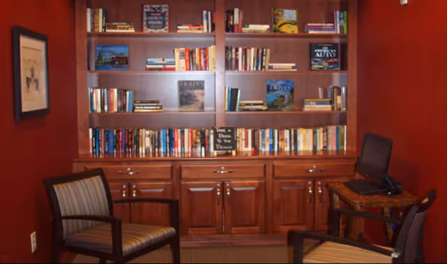 Cozy reading room with built-in wooden bookshelves filled with books, two upholstered chairs, and a small desk with a computer.