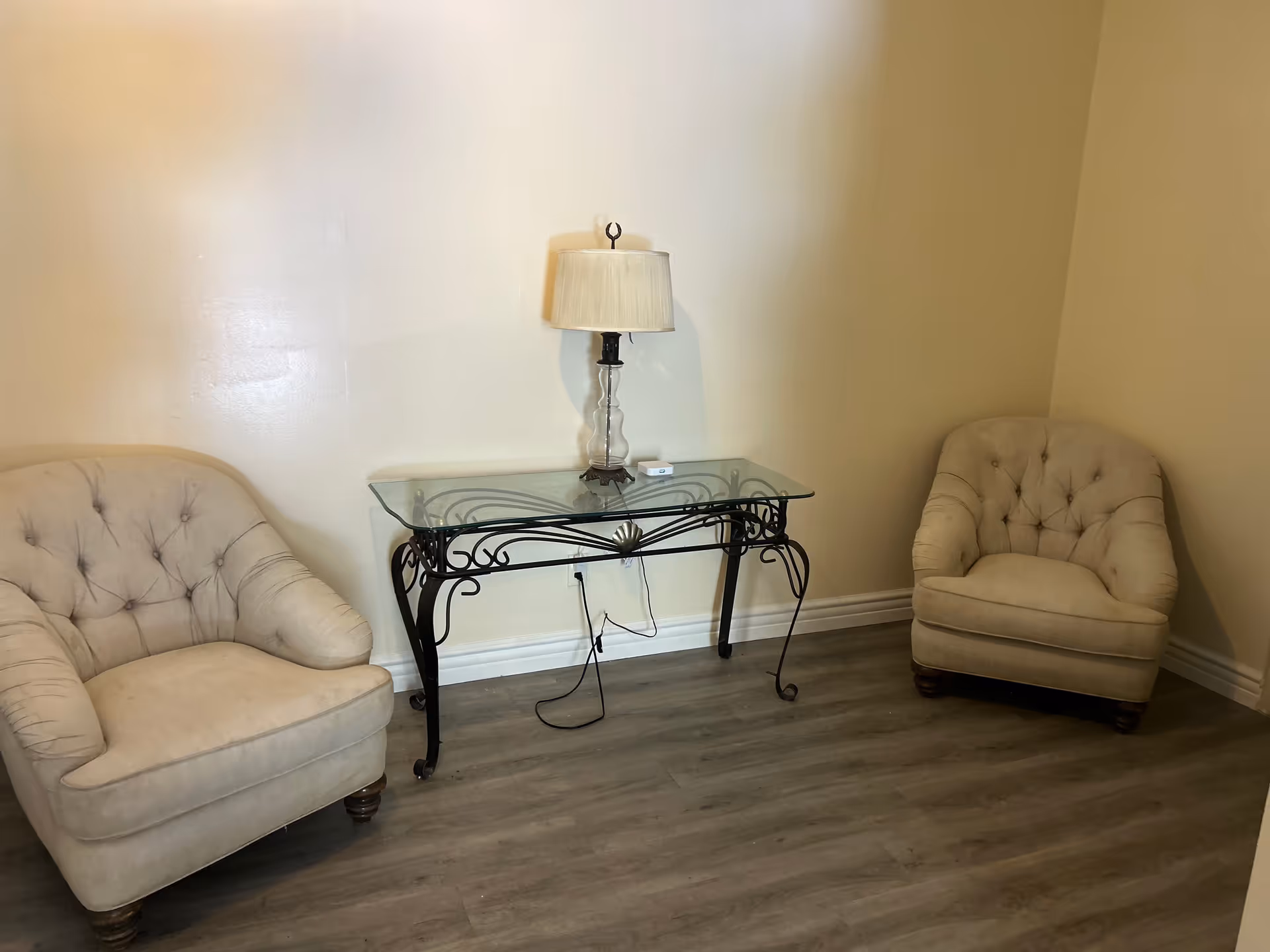 A small sitting area with two beige tufted armchairs placed on either side of a glass-top table with a decorative black metal frame. A table lamp with a beige shade is on the table. The floor is wood, and the walls are plain and light-colored.